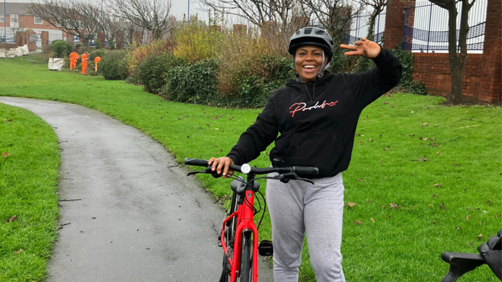 A woman stood with her bike and a helmet on smiling and doing a peace symbol with her hand