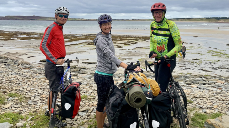 Three adults stood in cycling gear with their bikes and paniers smiling on the coast of Scotland