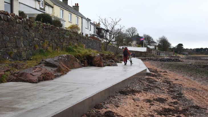 A person walks along the newly opened Fairlie Coastal Path pushing their bike. The tide is out, showing the beach and houses on either side of the traffic-free route.