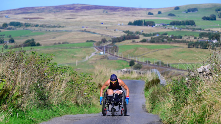 A woman in a wheelchair with her head down wheeling up a steep hill in the countryside on a sunny day