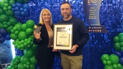 A woman holding a trophy and a man holding a certificate stand in front of a blue shiny wall surrounded by blue and green balloons.