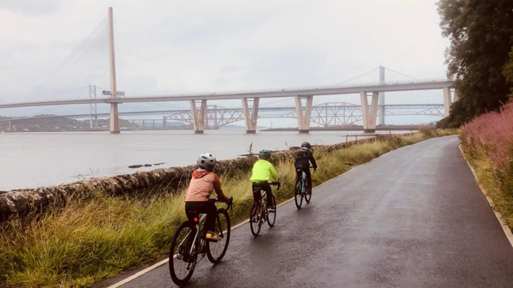 Three young boys on their bikes cycling on a road next to a body of water with bridges in the distance on an overcast day in Scotland