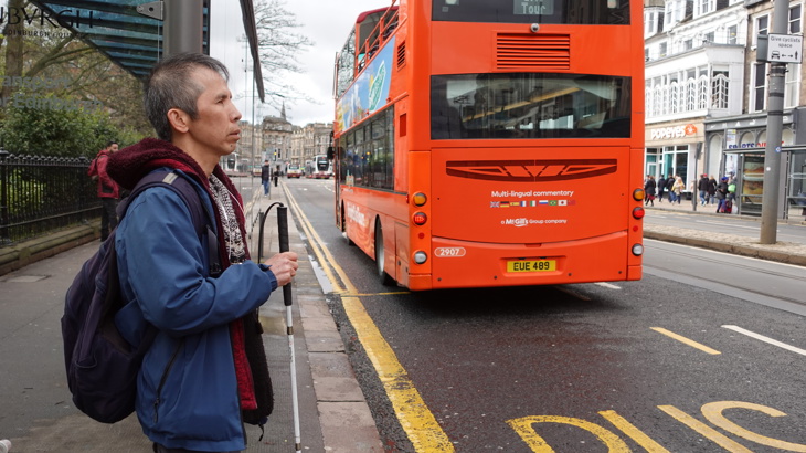 A man with a white cane waits at a bus stop as an orange double-decker bus pulls away on a city street.