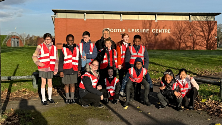 A group of primary school children wearing orange high-vis vests stood outside smiling in the sunshine