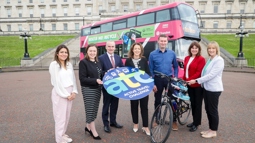 Five women and two men stand in front of a pink double decker bus parked in front of Stormont. They are holding a round blue sign saying Active Travel Challenge and there is also a bicycle with a pair of trainers hanging from the handlebars.
