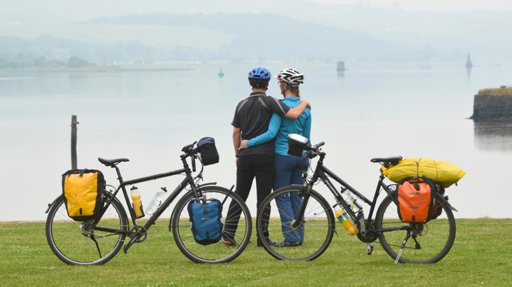 Two people standing together with their bikes, facing towards a body of water.