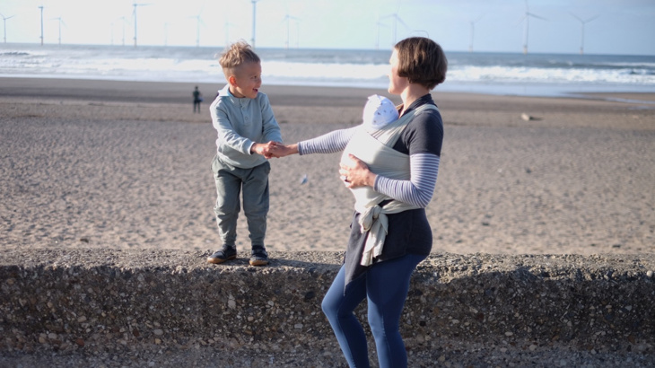 A woman and a newborn baby in a sling stood on a sea front on a bright day the woman is holding the hand of her small son who is stood up on a wall