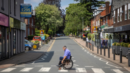 A man with white short white hair in a manual wheelchair crossing a zebra crossing on a quiet street in the UK on a sunny day