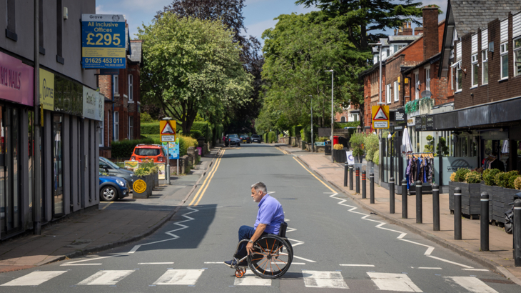 A man with white short white hair in a manual wheelchair crossing a zebra crossing on a quiet street in the UK on a sunny day