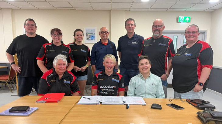 A group of people smiling at the camera with three people sat at a desk and the others stood up 