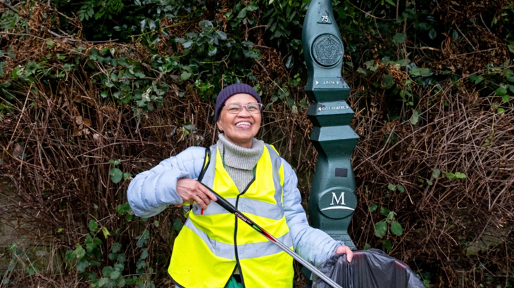 A woman in high-vis standing in front of a milepost on the Network smiling while litter picking 