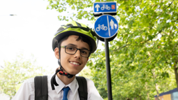 James smiles at the camera, wearing a yellow cycle helmet and a rucksack over a school shirt. Bright green trees and a blue cycling walking sign are in the background.