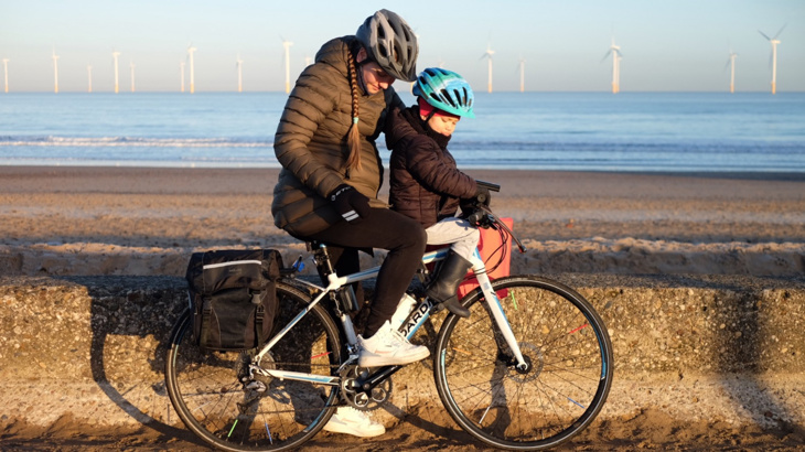 A mother stood on a bike with her toddler seated on the front, both wearing helmets and puffer jackets looking down at the woman's pregnancy bump on a sunny day on the coast in the East of England